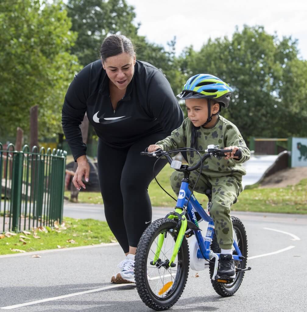 A mother encourages her little boy cycling unassisted on his bike at the children's cycle park