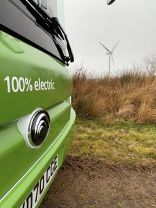 An electric bus front with the words '100% electric' on the bonnet, with a wind turbine in the background