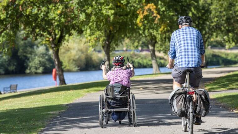 Two cyclists - one using an adapted bike cycling along the embankment